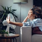 Eine Person sitzt auf einem Sofa vor einem Ventilator und h&auml;lt diesen fest, damit er nicht davonfliegt.