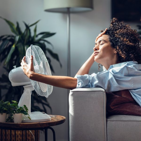 Eine Person sitzt auf einem Sofa vor einem Ventilator und h&auml;lt diesen fest, damit er nicht davonfliegt.