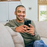 Eine Person sitzt auf der Couch im Wohnzimmer und scrollt auf ihrem Smartphone.