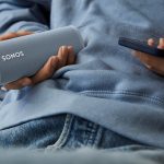 Eine Person h&auml;lt einen Sonos-Lautsprecher in der einen, ein Smartphone in der anderen Hand.