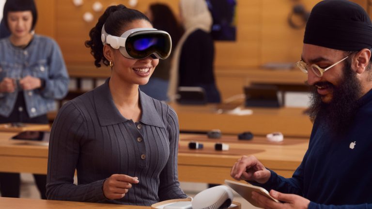 Eine Person hat in einem Apple Store die Vision Pro auf dem Kopf. Daneben steht ein*e Apple-Mitarbeiter*in mit einem iPad in der Hand.