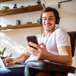 Eine Person sitzt auf einem Sessel, tr&auml;gt Kopfh&ouml;rer auf dem Kopf und bedient einen Laptop und ein Smartphone gleichzeitig.