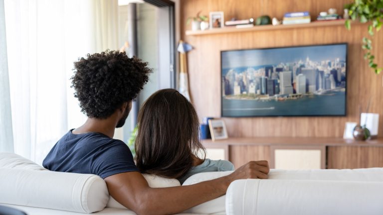 Ein P&auml;rchen sitzt im Wohnzimmer auf der Couch und sieht auf einem Fernseher fern.&nbsp;