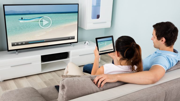 Ein P&auml;rchen sitzt im Wohnzimmer auf der Couch und &uuml;bertr&auml;gt ein Video von einem Tablet auf einen Fernseher.