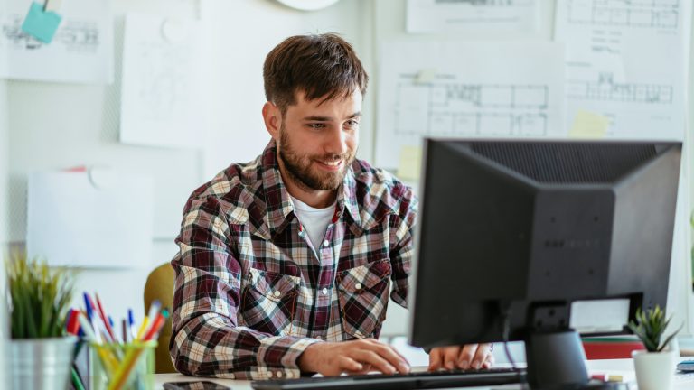 Eine Person sitzt l&auml;chelnd in einem B&uuml;ro vor einem Rechner.