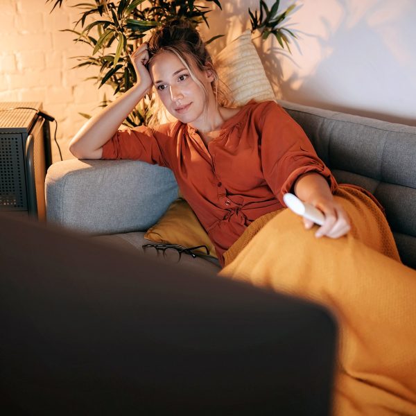 Eine Person liegt auf einem Sofa vor einem Fernseher und h&auml;lt eine Fernbedienung in der Hand.