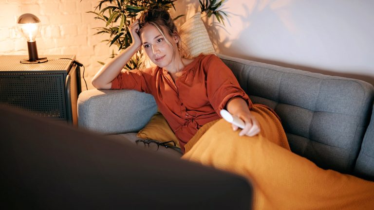 Eine Person liegt auf einem Sofa vor einem Fernseher und h&auml;lt eine Fernbedienung in der Hand.