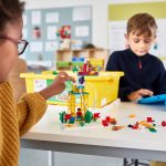 Zwei Kinder spielen an einem Tisch mit LEGO Spike. Eines davon bedient dabei ein Tablet.