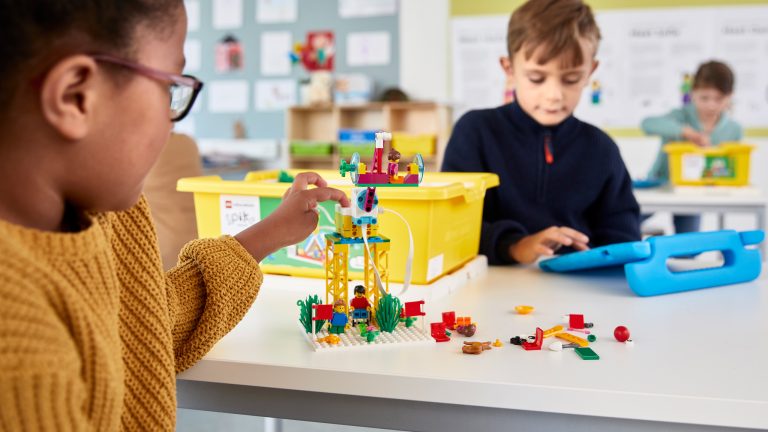 Zwei Kinder spielen an einem Tisch mit LEGO Spike. Eines davon bedient dabei ein Tablet.