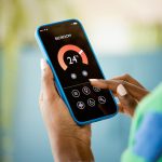 Eine Person h&auml;lt ein Smartphone in der Hand, das eine Temperaturanzeige und Einstellungsm&ouml;glichkeiten f&uuml;r ein Schlafzimmer anzeigt.