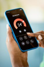 Eine Person h&auml;lt ein Smartphone in der Hand, das eine Temperaturanzeige und Einstellungsm&ouml;glichkeiten f&uuml;r ein Schlafzimmer anzeigt.