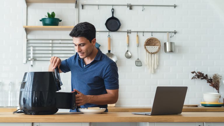 Eine Person steht in der Küche und holt etwas aus einem Airfryer. Daneben steht auf der Küche ein Laptop.