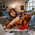 Eine Person liegt auf der Couch im Wohnzimmer mit Pflanzen im Hintergrund und scrollt auf ihrem Smartphone.