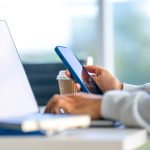 Eine Person h&auml;lt ein iPhone in der Hand und scheint an einem Schreibtisch zu sitzen.