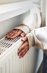 Heizung entlüften, In wenigen Schritten selbst gemacht Eine Person hält beide Hände an einen Heizkörper, der an einer Wand angebracht ist.