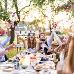 Eine Gruppe Personen sitzt mit einem Hochzeitspaar an einem langen Tisch und schmeißt Konfetti in die Luft.