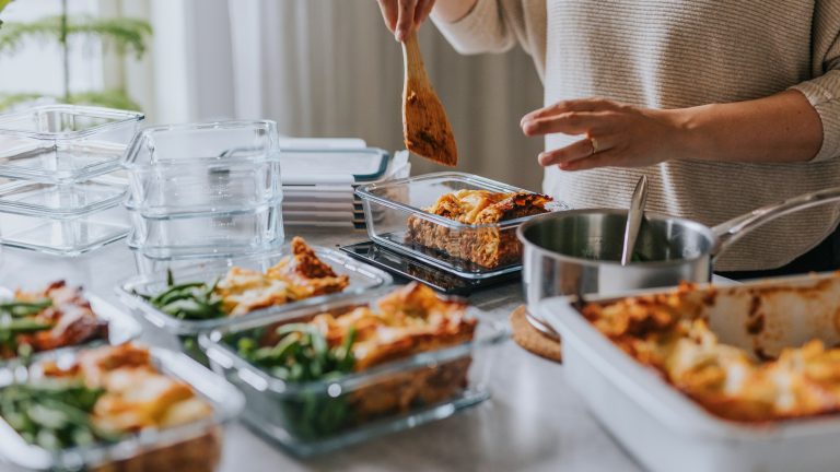 Eine Person befüllt beim MealPrepping auf einer Anrichte mehrere Glasboxen mit Lasagne und Bohnen.