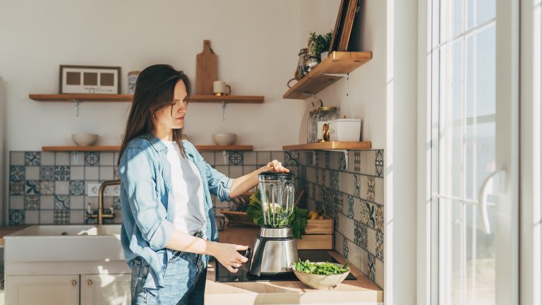 Eine Person steht in einer minimalistischen Küche und bedient einen Hochleistungsmixer, der auf der Küchenanrichte steht.