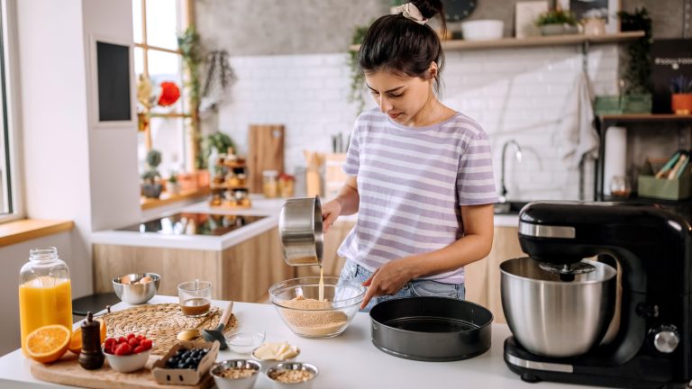 Eine Person gießt eine Flüssigkeit in eine Schüssel mit Mehl. Daneben stehen weitere Zutaten, wie Beeren und Eier, eine Backform und eine Küchenmaschine.