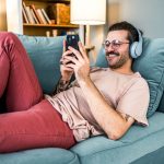Eine Person liegt auf einem Sofa, tr&auml;gt Kopfh&ouml;rer und h&auml;lt ein Smartphone in der Hand.