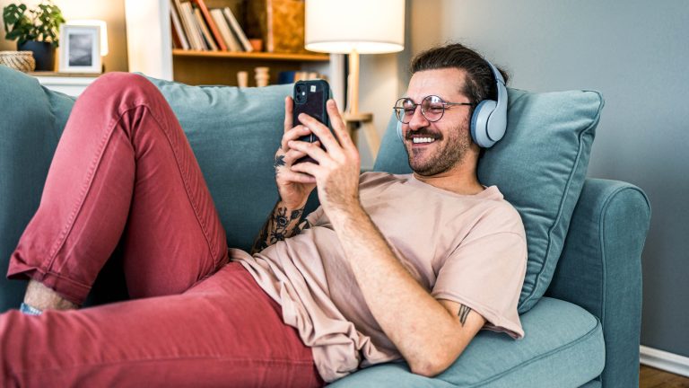Eine Person liegt auf einem Sofa, tr&auml;gt Kopfh&ouml;rer und h&auml;lt ein Smartphone in der Hand.
