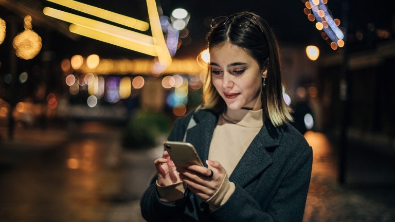 Eine Person steht nachts auf der Stra&szlig;e und blickt auf ihr Smartphone.