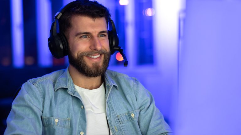 Eine Person mit einem Headset auf dem Kopf sitzt vor einem Rechner und grinst.