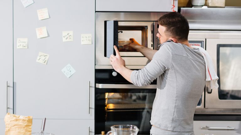 Eine Person stellt eine Sch&uuml;ssel mit Essen in die Mikrowelle w&auml;hrend sie nebenbei telefoniert.