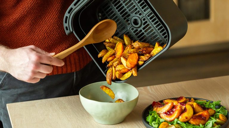 Eine Person gie&szlig;t einige Kartoffelspalten von einem Airfryer-Korb in eine Sch&uuml;ssel.