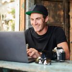 Eine Person mit einer Kamera neben ihr auf dem Tisch sitzt an einem Laptop und grinst.