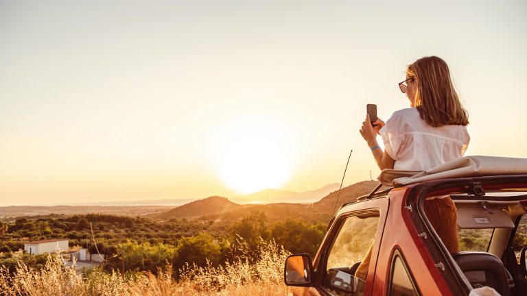 Eine Person steht in einem Auto und fotografiert eine Landschaft und den Sonnenuntergang durch das offene Verdeck.
