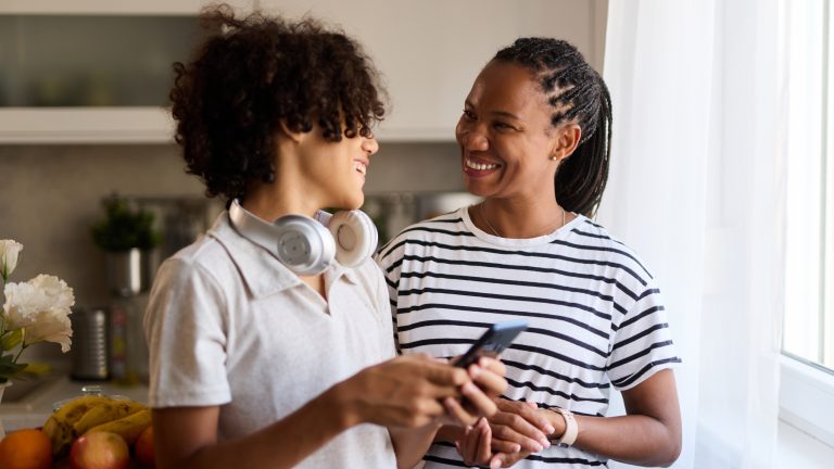 Eine erwachsene und eine jugendliche Person stehen nebeneinander und lachen sich an. Die jugendliche Person h&auml;lt ein Smartphone in der Hand.
