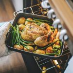 Eine Person steckt eine Auflaufform mit H&auml;hnchen, Kartoffeln und gr&uuml;nen Bohnen in einen ge&ouml;ffneten Backofen.