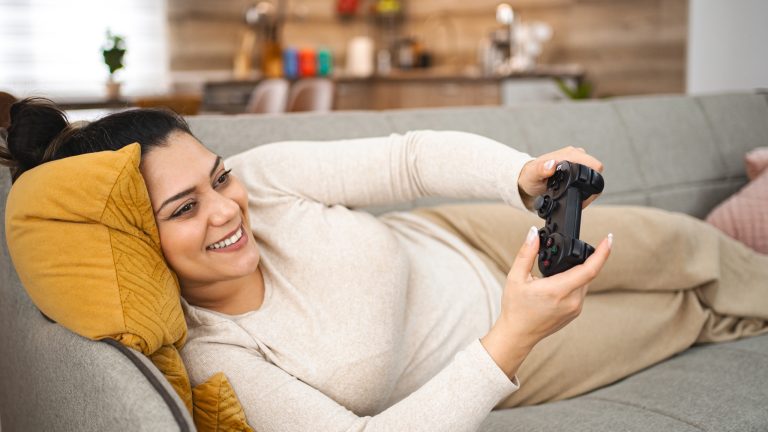 Eine Person liegt gem&uuml;tlich auf einem Sofa und h&auml;lt einen Controller in der Hand.