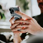 Ein iPhone in den H&auml;nden einer Person, die auf einer Couch vor einem Fenster sitzt.
