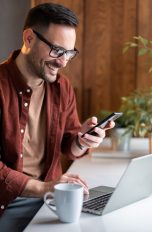 Passkey, Was sie sind und wie sie funktionieren Eine Person sitzt mit einem Kaffee vor einem Laptop und hält ein Smartphone in der Hand.