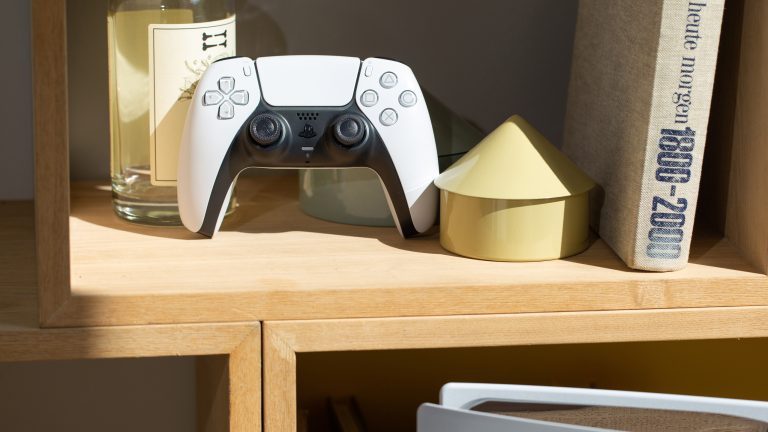 Ein Dualsense-Controller steht in einem Regal. Im Vordergrund ist eine PS5 zu erkennen.