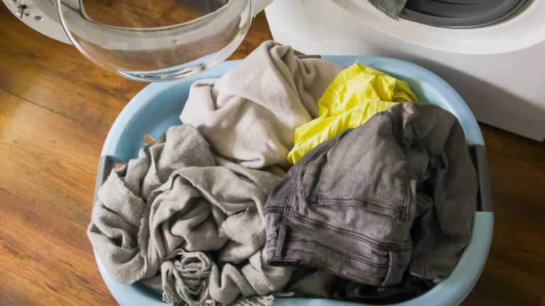 Waschmaschinen-Symbole NEU Eine offene Waschmaschine, in der noch etwas Wäsche steckt. Davor steht ein Korb mit weiterer Wäsche.