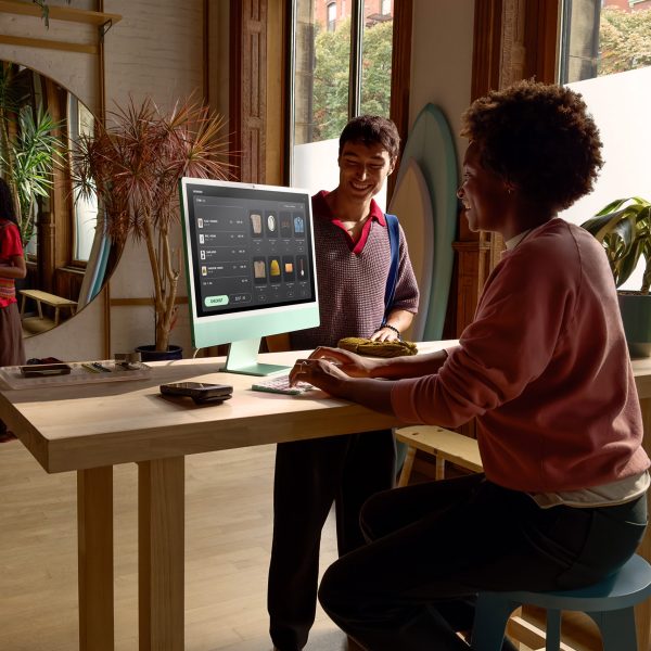 Eine Person sitzt vor einem iMac 2024. Ihr gegen&uuml;ber steht eine weitere Person.