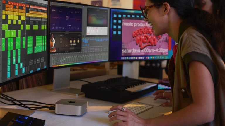 Zwei Personen stehen vor einem Mac mini, an den drei Monitor angeschlossen sind. Sie scheinen Musik zu produzieren.