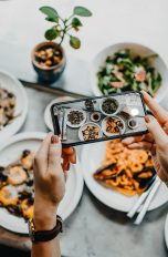 Food Fotografie, Tipps und Tricks um leckeres Essen gut zu inszenieren Nahaufnahme der Hände einer Person, die mit dem Smartphone Fotos von einem gedeckten Tisch macht.