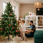 Zwei Personen sitzen auf dem Boden im Wohnzimmer, w&auml;hrend ein Kind den Weihnachtsbaum schm&uuml;ckt.