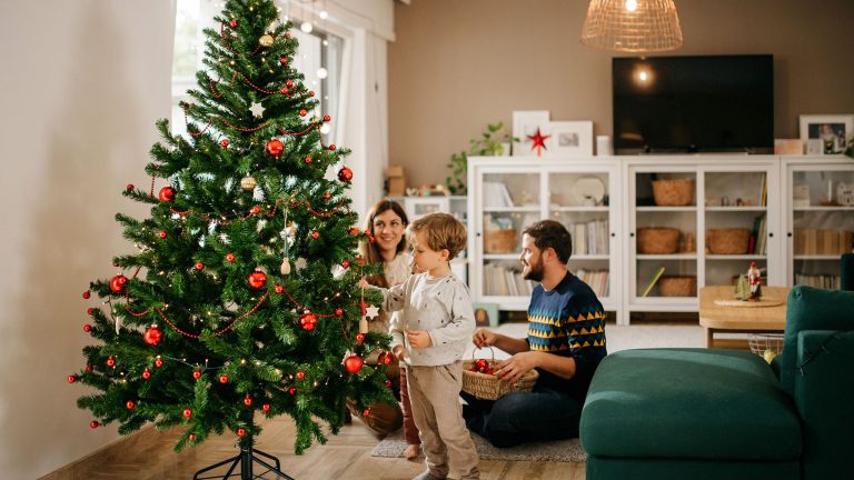 Zwei Personen sitzen auf dem Boden im Wohnzimmer, w&auml;hrend ein Kind den Weihnachtsbaum schm&uuml;ckt.