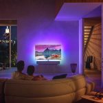 R&uuml;ckansicht zweier Personen, die Arm in Arm auf einer Couch sitzen und eine Szene auf dem Fernseher ansehen, die die dahinterliegende Wand in lila-blaues Licht taucht.