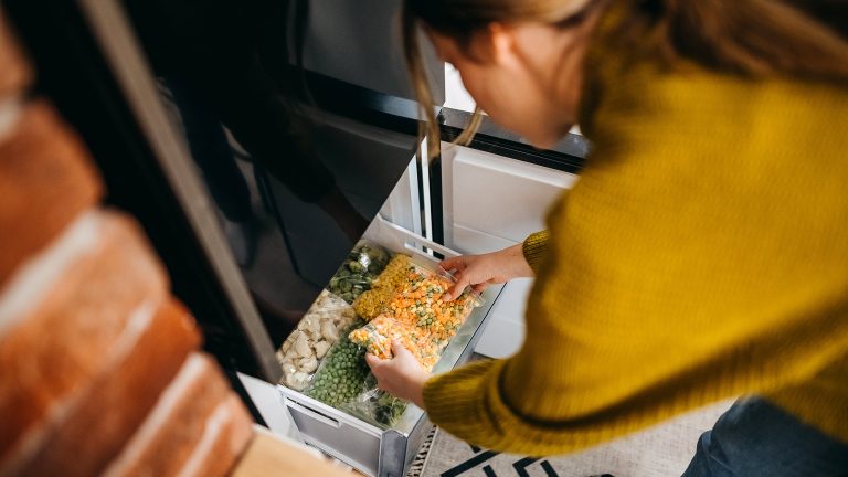 Eine Person legt eine durchsichtige Tüte mit buntem Gemüse in die Schublade eines Gefrierschrankes.