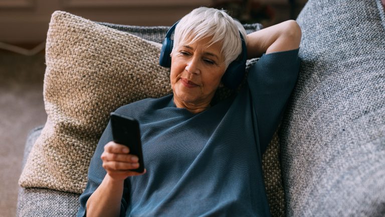 Eine Person liegt auf dem Sofa und h&ouml;rt &uuml;ber Kopfh&ouml;rer etwas an ihrem Handy auf das sie schaut.