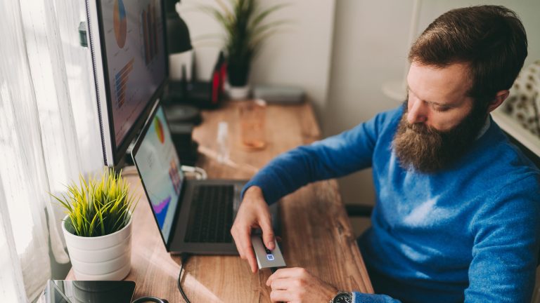 Eine Person sitzt am Tisch vor einem Laptop und versucht mithilfe eines USB-Anschlusses eine externe Festplatte zu verbinden.