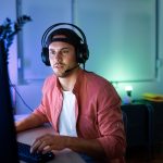 Eine Person sitzt mit einem Headset vor einem PC &ndash; H&auml;nde an der Maus und der Tastatur. Der Raum ist blau und gr&uuml;n beleuchtet.