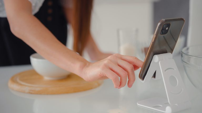 Ein Smartphone steht auf einem Standfu&szlig; auf einer Arbeitsfl&auml;che in einer K&uuml;che. Im Hintergrund ist eine wei&szlig;e Sch&uuml;ssel auf einem Holzbrett zu sehen.