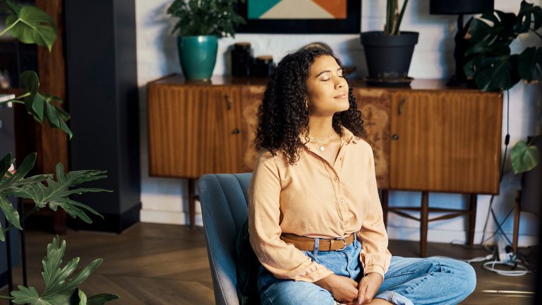Eine Person sitzt mit geschlossenen Augen auf einem Stuhl. Das Gesicht streckt sie der Sonne entgegen, die durch das Fenster scheint.